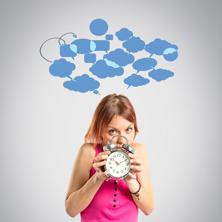 Serious redhead girl holding a clock over grey background の写真素材