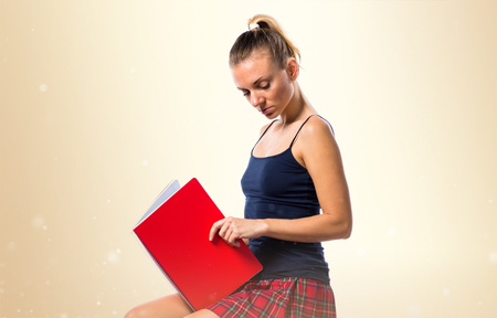 Pretty blonde school girl sitting on many books and holding notebooks on ocher backgroundの写真素材