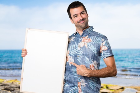 Handsome man with flower shirt holding an empty placard at the beachの写真素材