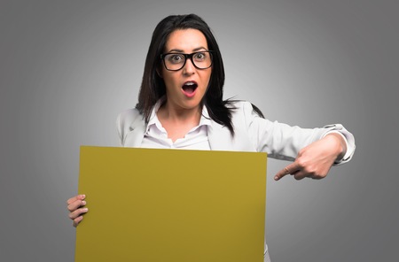 Pretty woman with glasses holding an empty placard on grey backgroundの写真素材