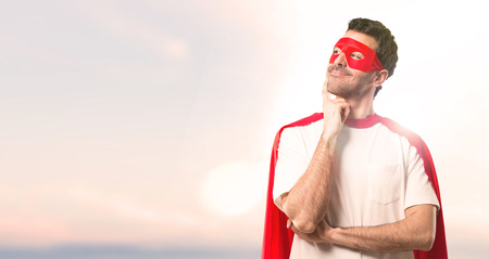Superhero man with mask and red cape standing and thinking an idea while looking up on a sunset backgroundの写真素材