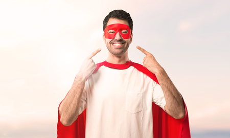 Superhero man with mask and red cape smiling with a happy and pleasant expression while pointing mouth and face with fingers on a sunset backgroundの写真素材