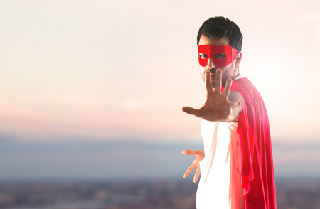 Superhero man with mask and red cape making stop sign on a sunset backgroundの写真素材