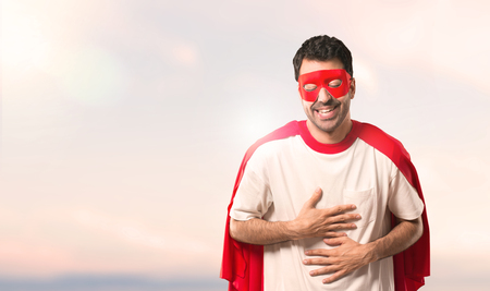 Superhero man with mask and red cape smiling a lot while putting hands on chest on a sunset backgroundの写真素材
