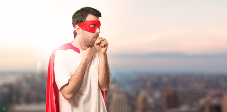 Superhero man with mask and red cape is suffering with cough and feeling bad on a sunset backgroundの写真素材