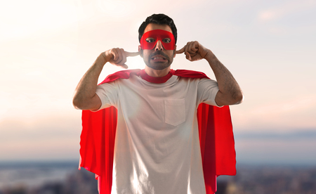 Superhero man with mask and red cape covering his ears on a sunset backgroundの写真素材
