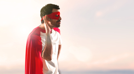 Superhero man with mask and red cape listening something on a sunset backgroundの写真素材