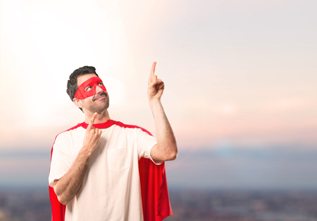 Superhero man with mask and red cape pointing with the index finger a great idea and looking up on a sunset backgroundの写真素材