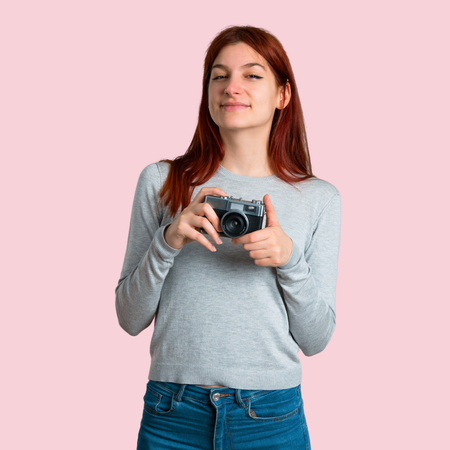 Young redhead girl holding a camera on isolated pink backgroundの写真素材