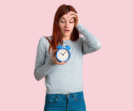 Young redhead girl restless because it has become late and holding vintage alarm clock on isolated...