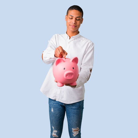 Young african american man with white shirt taking a piggy bank and happy because it is full on isolated blue backgroundの写真素材