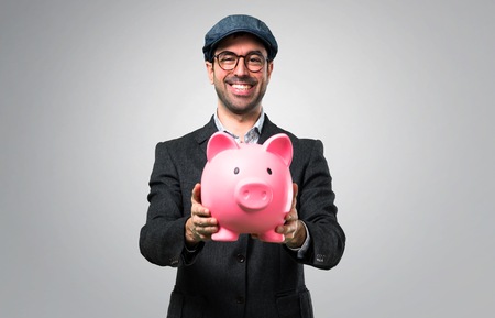 Handsome modern man with beret and glasses holding a piggybank on grey backgroundの写真素材