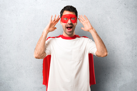 Superhero man with mask and red cape with surprise and shocked facial expression. Gaping because can not believe what is happening on textured grey backgroundの写真素材