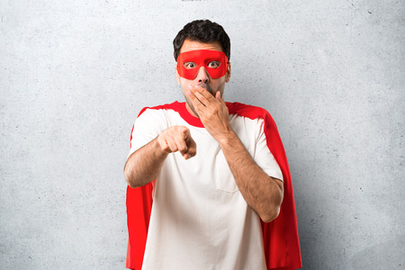 Superhero man with mask and red cape pointing with finger at someone and laughing a lot while covering mouth on textured grey backgroundの写真素材