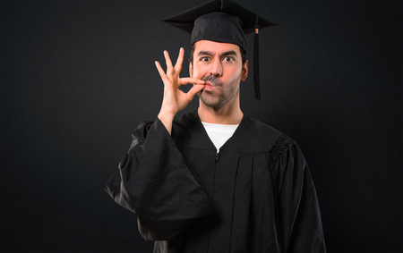 Man on his graduation day University showing a sign of closing mouth and silence gesture doing like closing his mouth with a zipper on black backgroundの写真素材