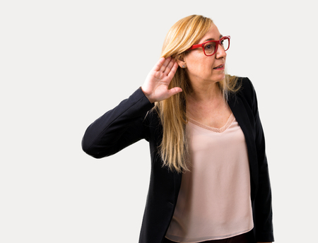 Middle-age blonde business woman with black jacket listening to something by putting hand on the ear on isolated grey backgroundの写真素材