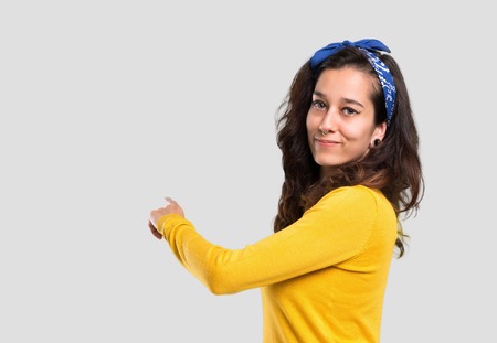 Young girl with yellow sweater and blue bandana on her head pointing back with the index finger presenting a product from behind on isolated grey backgroundの写真素材