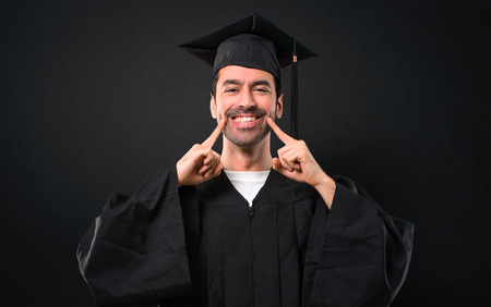 Man on his graduation day University smiling with a happy and pleasant expression while pointing mouth and face with fingers on black backgroundの写真素材