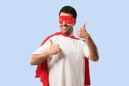 Superhero man with mask and red cape giving a thumbs up gesture and smiling because has had success on isolated blue backgroundの写真素材