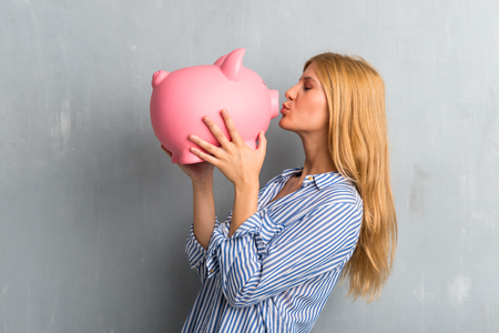 Blonde girl holding a big piggybank on textured grunge wall backgroundの写真素材