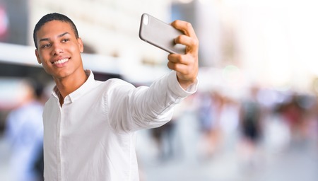 Young african american man with white shirt taking a selfie with the mobile in the middle of the cityの写真素材