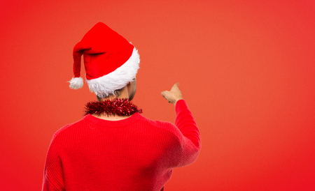 Man with red clothes celebrating the Christmas holidays pointing back with the index finger on red backgroundの写真素材
