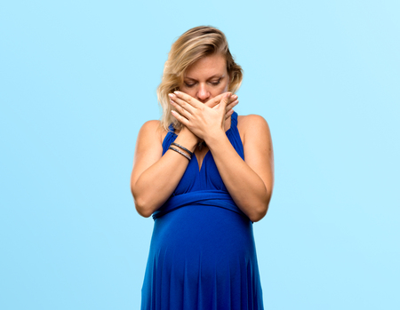 Pregnant blonde woman with blue dress covering mouth with hands on isolated blue backgroundの写真素材