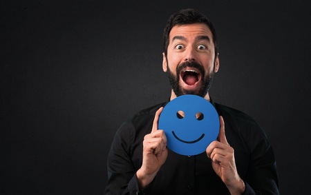 Handsome man with beard holding a happy face icon on black backgroundの写真素材
