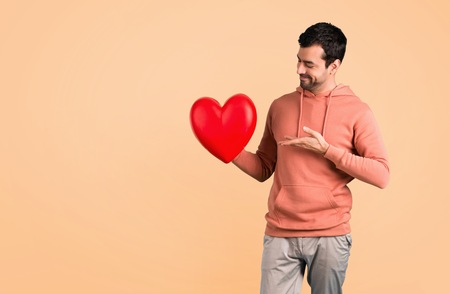 Man in a pink sweatshirt holding a big heart icon toy on ocher background