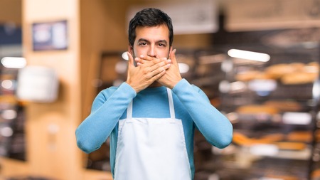 Man wearing an apron covering mouth with hands for saying something inappropriate. Can not speak in a bakeryの写真素材