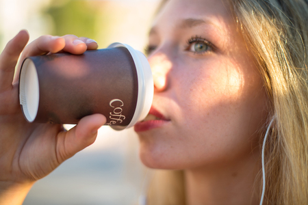 Young blonde girl listening to music with headphones and drinking hot coffee in takeaway paper cup