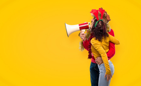 Couple dressed up for the christmas holidays holding a megaphone on yellow background