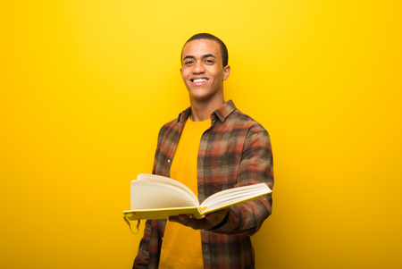 Young african american man on vibrant yellow background holding a book and giving it to someoneの写真素材