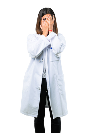 Doctor woman with stethoscope covering eyes by hands and looking through the fingers on isolated white backgroundの写真素材