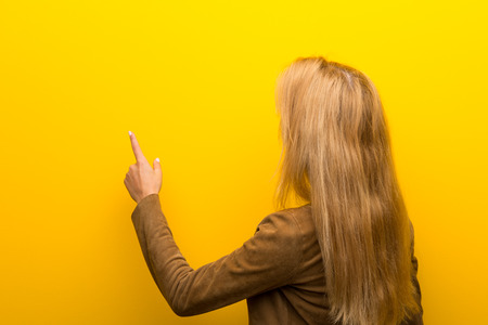 Blonde girl on vibrant yellow background pointing back with the index fingerの写真素材