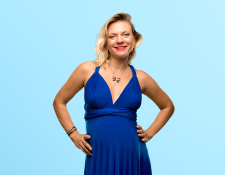 Pregnant blonde woman with blue dress posing with arms at hip on isolated blue backgroundの写真素材