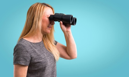 Middle-age blonde woman and looking for something in the distance with binoculars on colorful backgroundの写真素材