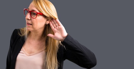 Middle-age blonde business woman with black jacket listening to something by putting hand on the ear...