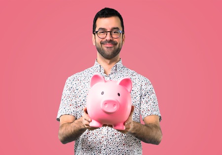 Handsome man with glasses holding a piggybank on pink backgroundの写真素材