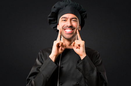 Chef man In black uniform smiling with a happy and pleasant expression while pointing mouth and face with fingers on black backgroundの写真素材