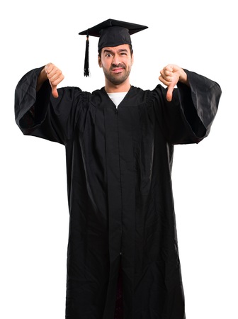 Man on his graduation day University showing thumb down with both hands. Negative expression on isolated white backgroundの写真素材