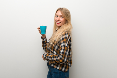 Blonde young girl over white wall holding a hot cup of coffeeの写真素材