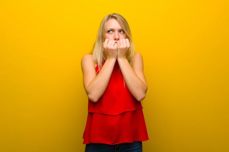 Young girl with red dress over yellow wall is a little bit nervous and scared putting hands to mouthの写真素材