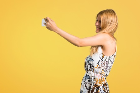 Blonde young girl in a summer dress taking a selfie with the mobile on yellow backgroundの写真素材