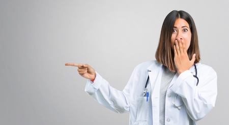 Doctor woman with stethoscope pointing finger to the side and presenting a product with a surprised face while covering mouth on grey backgroundの写真素材