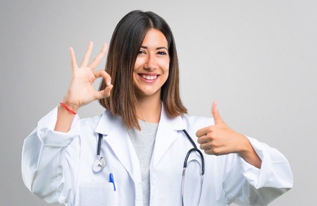 Doctor woman with stethoscope showing an ok sign with fingers and giving a thumb up gesture with the other hand on grey backgroundの写真素材