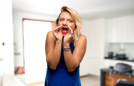 Pregnant blonde woman with blue dress shouting with mouth wide open in her houseの写真素材