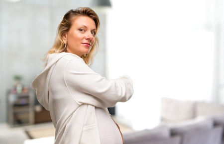 Pregnant blonde woman with white sweatshirt pointing back with the index finger in her houseの写真素材