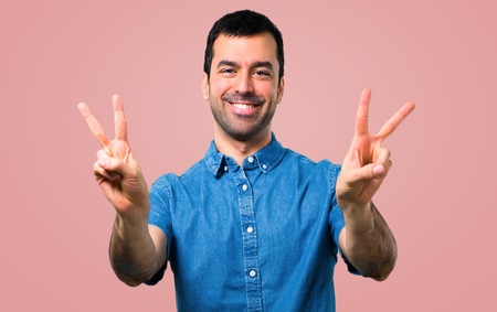 Handsome man with blue shirt smiling and showing victory sign on pink background