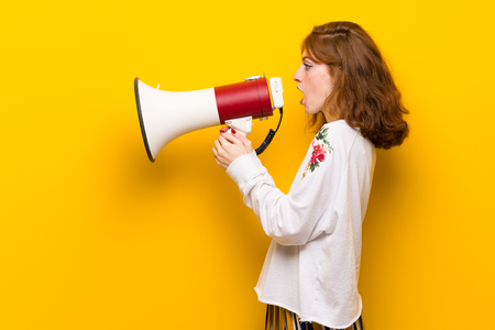 Young redhead woman over yellow wall shouting through a megaphone to announce something in lateral positionの写真素材